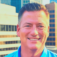Smiling man in a blue polo shirt with a blurred city skyline in the background.